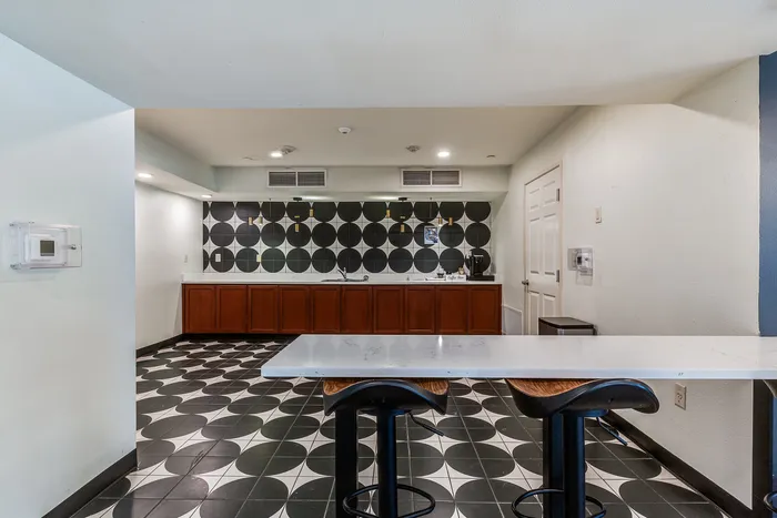 Community kitchen and lounge area at Onyx at Oslo Apartments in San Antonio Texas showing a long counter with wooden cabinets, black and white geometric wall tiles, pendant lights, a coffee station, a sink, black and white patterned floor tiles, and a high table with bar stools in the foreground.