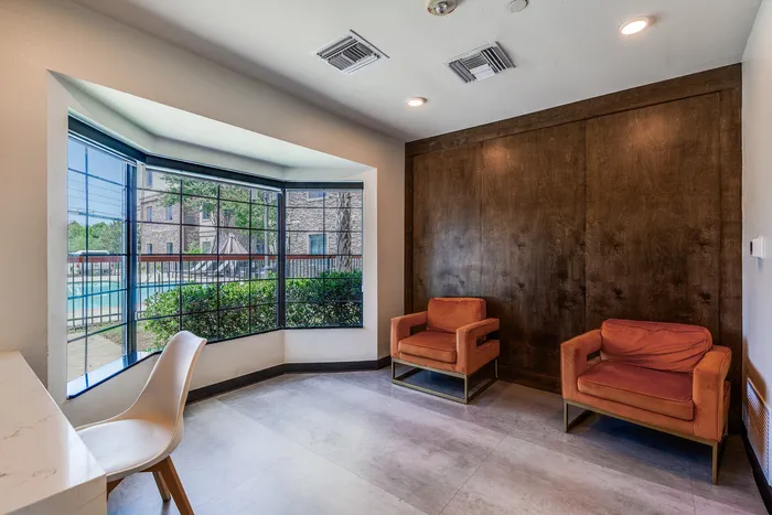 Small seating area at Onyx at Oslo Apartments in San Antonio Texas with two orange armchairs against a dark wood wall, a white desk with a chair, and a large bay window looking out toward the pool and exterior buildings.