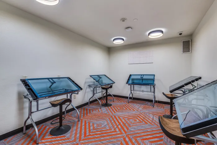 Art room at Onyx at Oslo Apartments in San Antonio Texas with several adjustable glass top drafting tables, stools, white walls, and patterned orange and gray flooring.
