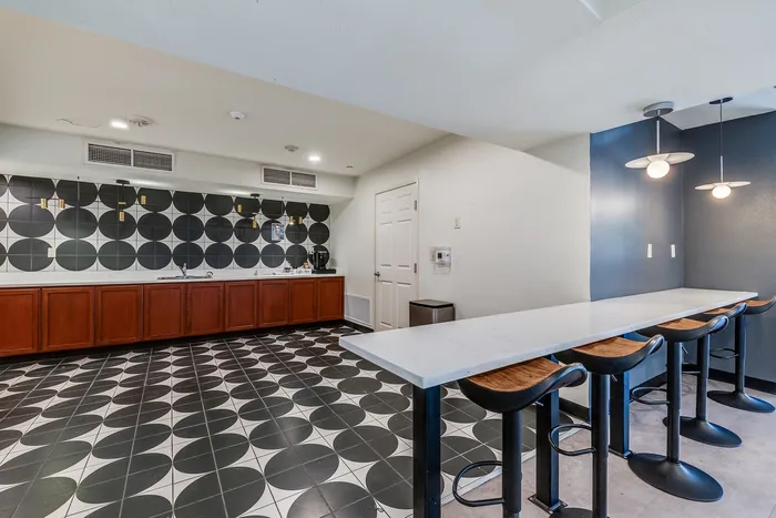 Coffee bar area at Onyx at Oslo with a long white counter, wooden bar stools, pendant lights, and a full counter wall with black and white circular tile backsplash.