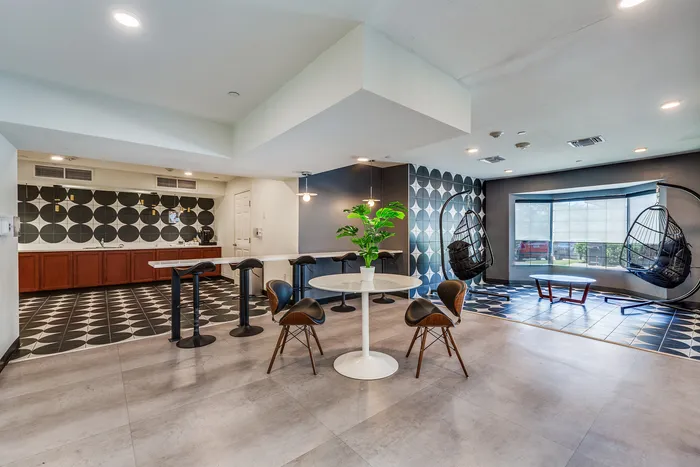 Coffee bar area at Onyx at Oslo with a long counter and bar stools, round table with chairs, patterned tile accent wall, and two hanging basket chairs near a large window.