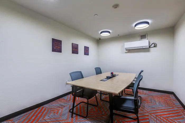 A small conference room with a wooden meeting table, four black chairs, bright overhead lights, wall art, and patterned orange and gray carpet.