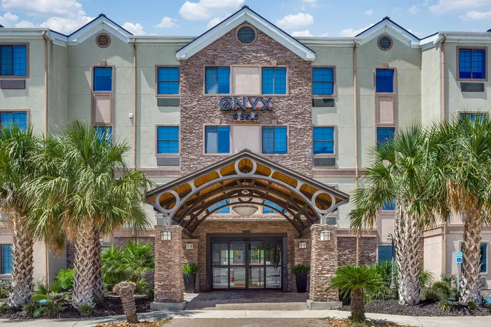 Front exterior of the Onyx at Oslo building with stone accents, blue windows, palm trees, and a covered entryway leading to glass doors.