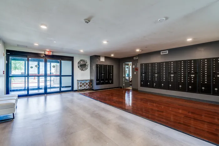 Bright lobby area with large glass doors, dark mailbox walls on both sides, a wood flooring section, and a decorative mirror near the mailboxes.