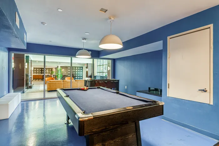 A blue game room with a pool table in the center, bright overhead lights, and glass doors opening into a lounge area.