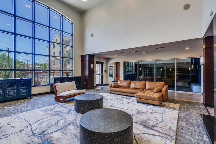 Lobby with tall windows, a leather sofa, lounge chair, and round tables in a bright common area.