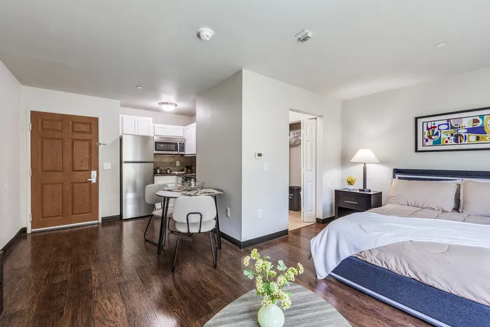 Open apartment layout showing a kitchen with stainless appliances, a small dining table, and a bedroom area with a nightstand and wall art.