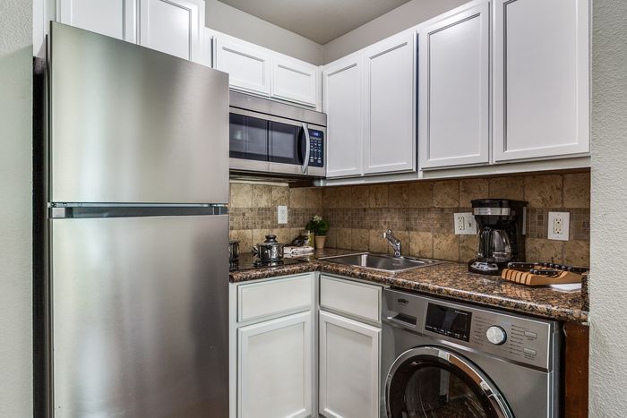 A modern apartment kitchen featuring white cabinets, a stainless refrigerator, microwave, sink, and in-unit washer and dryer, with granite countertops and tiled backsplash.