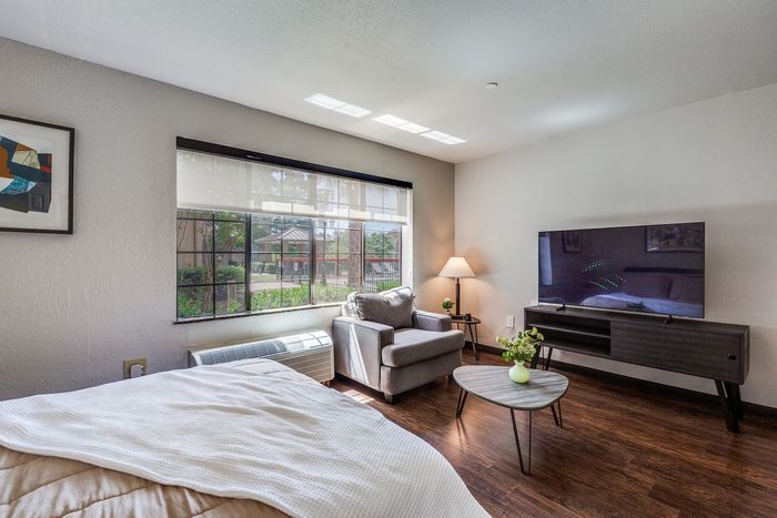 A bedroom setup with a seating corner, large window, and TV console, featuring wood-style flooring.