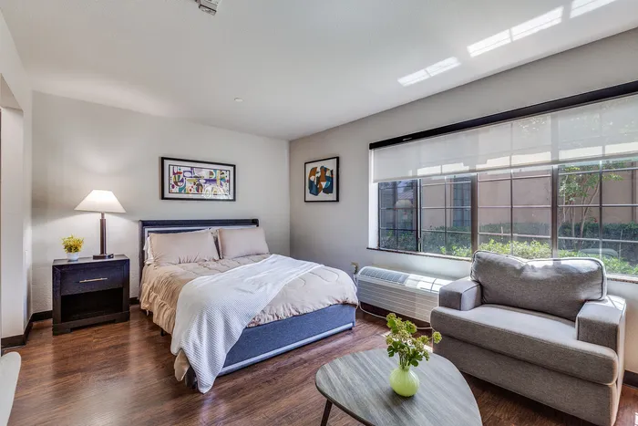 A bright bedroom layout with a seating area and large windows letting in natural light.