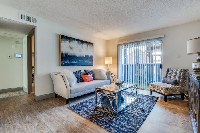 A spacious living area featuring wood-style flooring, large windows with vertical blinds, and access to a private patio.