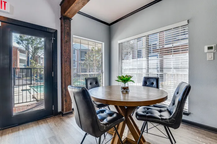 A small seating area with a round wooden table, four padded chairs, and large windows letting in natural light.