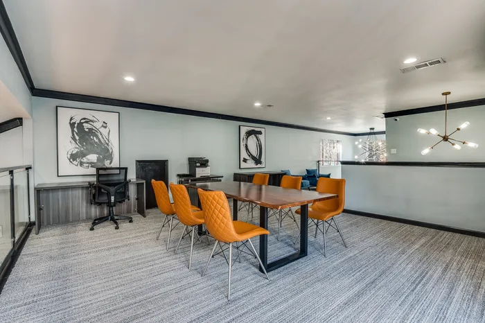 A room featuring a long wooden table with orange chairs, additional desk seating, modern lighting, and soft carpet flooring.