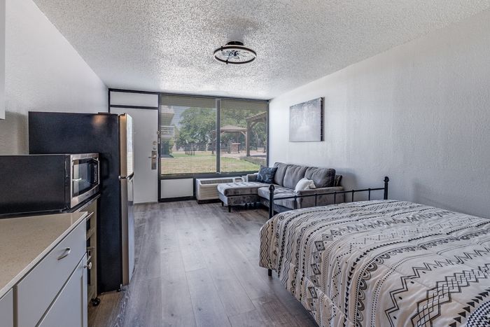 Bright studio apartment with elegant wood flooring, modern kitchen, cozy lounge area, and large windows offering a serene outdoor view.