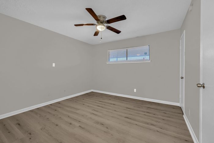 Modern bedroom with sleek wooden flooring and contemporary ceiling fan, offering a versatile space with natural light from horizontal blinds.