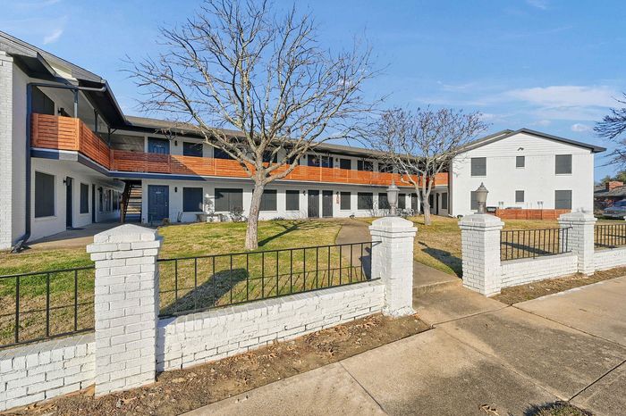 Charming two-story apartment building with modern wooden railings and a well-maintained lawn, framed by elegant white brick fencing.