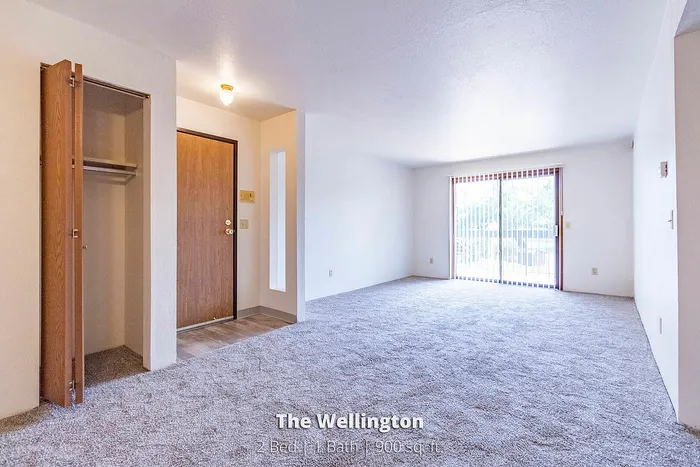 Bright and spacious living area in The Wellington apartment, featuring plush carpeting and large sliding glass doors leading to a private balcony. 2 Bed, 1 Bath, 900 sq ft.