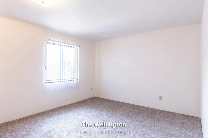 Bright, spacious room in The Wellington apartment, featuring a large window with vertical blinds, perfect for a cozy and inviting atmosphere. 2 bed, 1 bath, 900 sq ft.