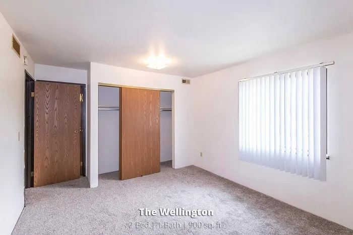 Bright and welcoming bedroom with soft carpet, ample closet space, and a large window at The Wellington apartments, featuring 2 bedrooms and 1 bath in 900 sq. ft.