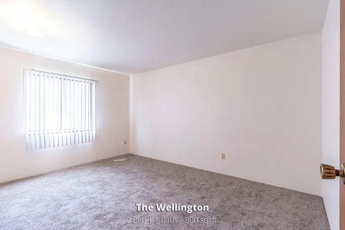 Inviting bedroom in The Wellington showcasing plush carpeting, ample natural light through vertical blinds, and spacious layout for cozy living.