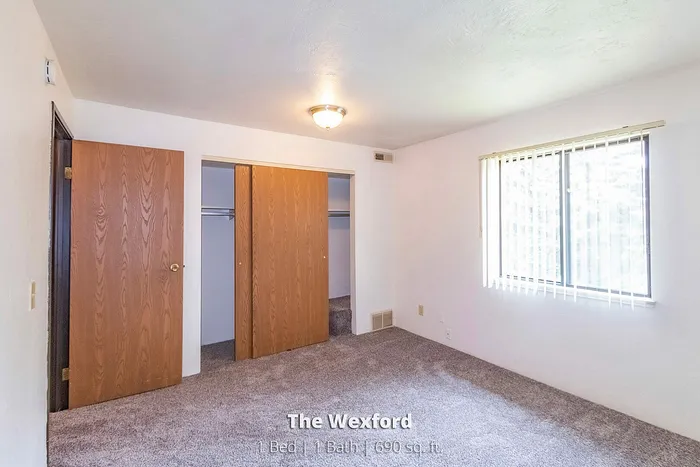 Bright and airy bedroom in The Wexford, featuring a spacious closet, large window with natural light, and plush carpet. Cozy 1 bed, 1 bath apartment with 690 sq. ft.