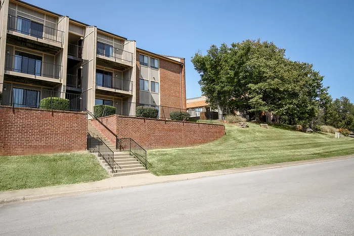 Charming three-story apartment building with private balconies, nestled on a grassy hillside with mature trees and a serene neighborhood street.