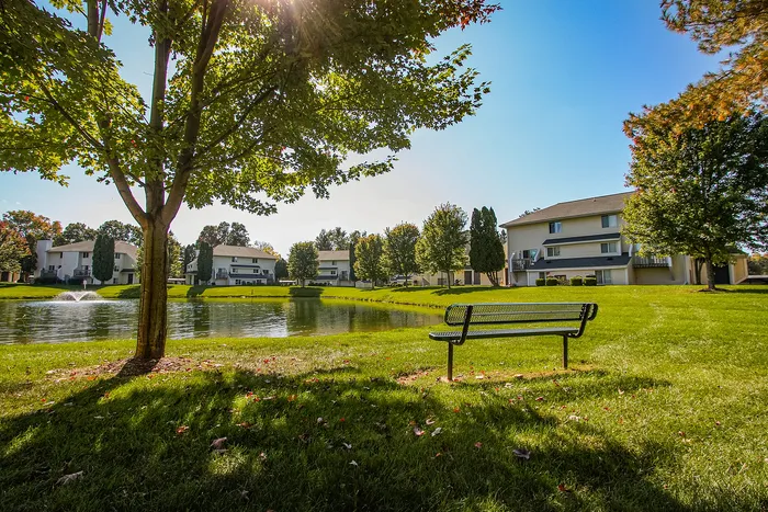 Serene apartment complex with nearby pond, surrounded by lush green lawns and mature trees, under a sunny blue sky.