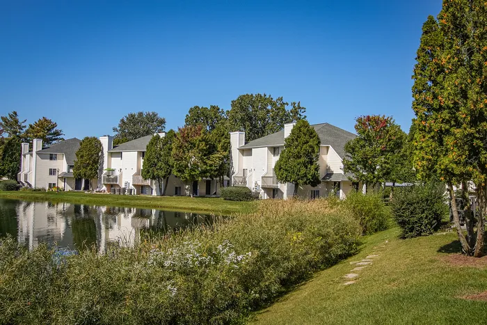 Charming lakeside apartments surrounded by lush greenery and vibrant trees under a clear blue sky, offering serene and picturesque living.