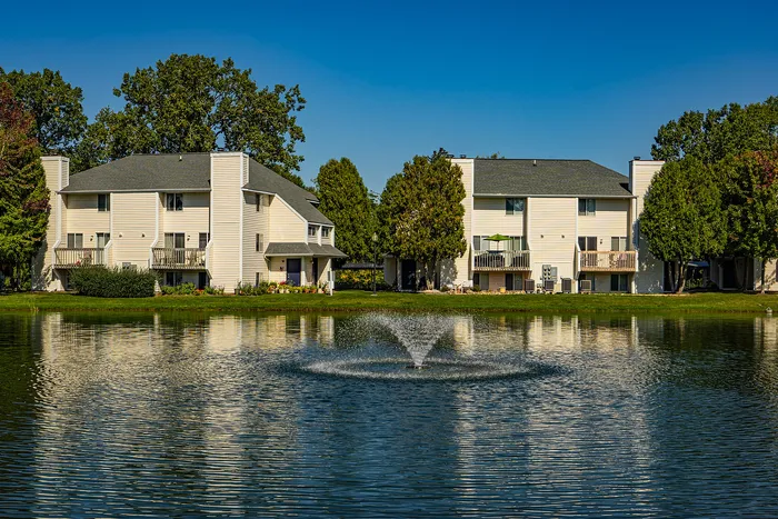 Serene apartment complex with modern white buildings and private balconies overlooking a tranquil pond with a fountain, surrounded by lush green trees.