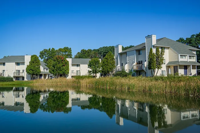 Serene lakeside apartment complex featuring modern white buildings with balconies, surrounded by lush greenery and reflected in tranquil water.