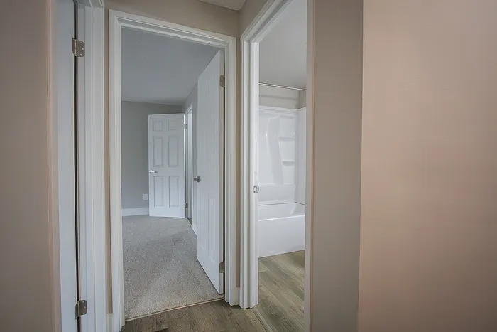 Inviting apartment hallway with wood flooring leading to a carpeted bedroom and modern bathroom, showcasing stylish white doors.