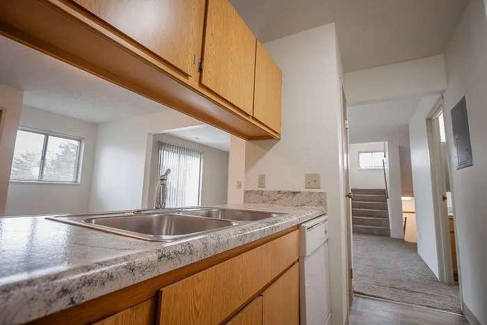 Bright and inviting kitchen with sleek marble countertops, stainless steel double sink, and elegant wooden cabinets, leading to a cozy carpeted stairway.