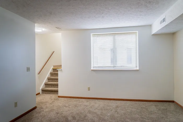 Charming apartment interior with plush carpet, stylish staircase, and bright natural light streaming through a large window.