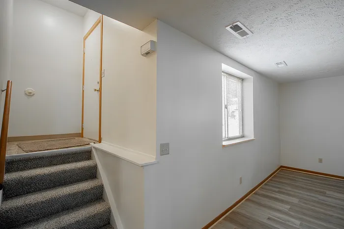 Inviting entrance with a carpeted staircase leading to a bright, modern room featuring a large window and sleek wood flooring.