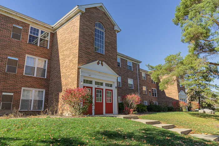 Charming brick apartment building with red entry doors, lush greenery, and a welcoming path on a sunny day.
