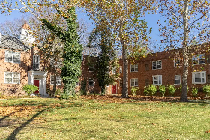 Charming brick apartment complex with red doors, nestled among towering trees and lush greenery, bathed in bright sunlight.