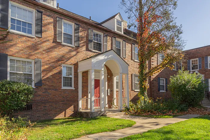 Charming brick apartment building with colonial architecture, featuring a welcoming red door, lush greenery, and sunlit facade.