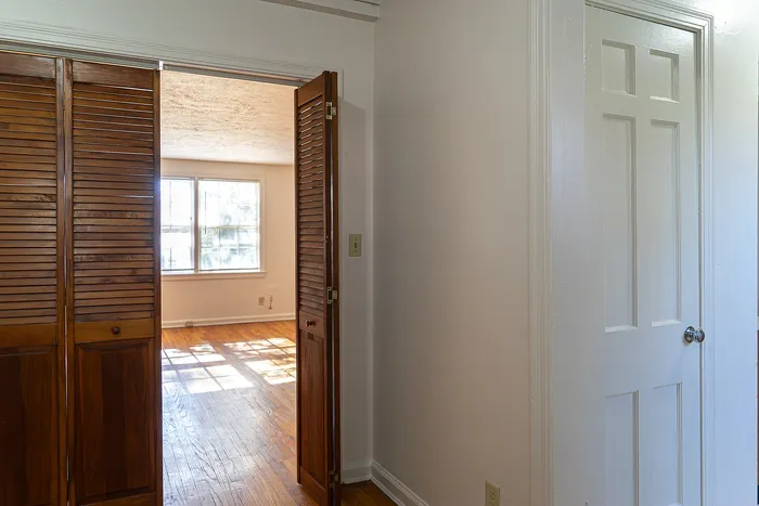 Inviting apartment interior with warm wooden floors, sunlight streaming through large windows, and charming wooden louvered doors.