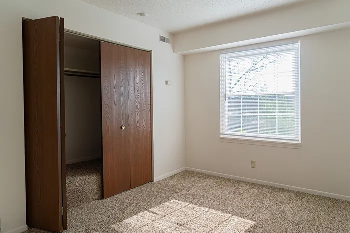 Bright, cozy bedroom with plush carpet, natural light from large window, and spacious wooden closet.