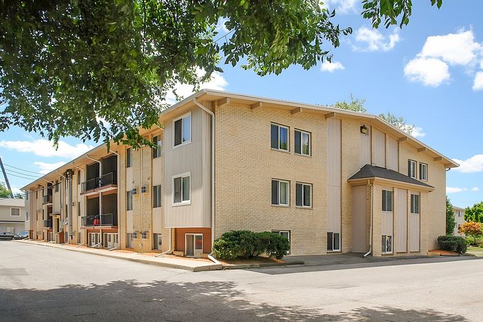 Charming three-story brick apartment building with lush greenery, offering sunlit windows and modern amenities.