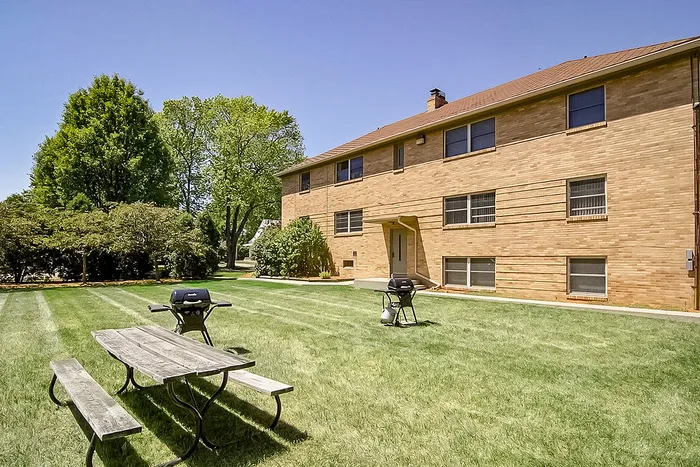 Charming red-brick apartment building with lush green lawn, picnic table, and barbecue grills, ideal for outdoor gatherings.