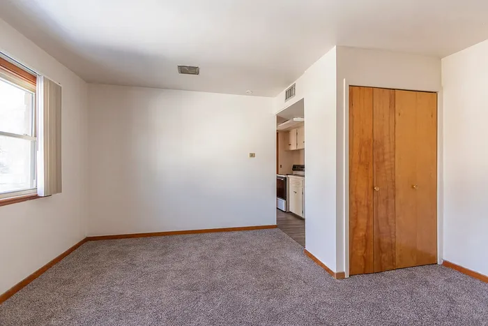 Bright and inviting living space with plush carpet, an open doorway leading to a modern kitchen, and large natural light-filled windows.