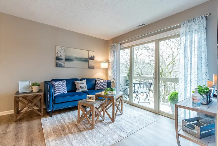 Bright and cozy living room with a modern blue sofa, wooden tables, and large glass doors opening to a scenic balcony.