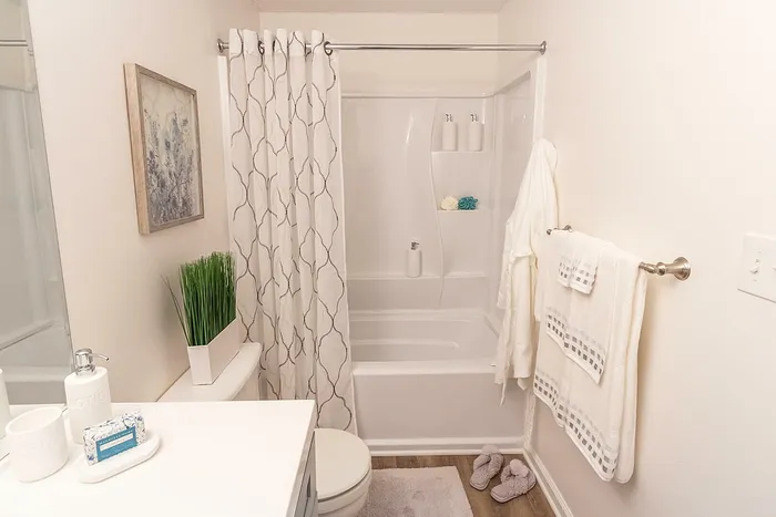 Elegant bathroom featuring a sleek tub with a white curtain, stylish wall art, and modern accessories. Plush towels and bath slippers add a cozy touch.