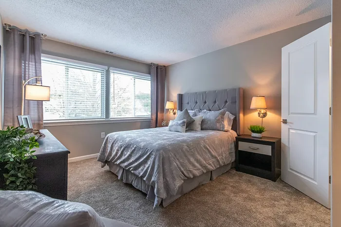 Inviting bedroom with plush gray décor, elegant bedside lighting, and abundant natural light from large windows.