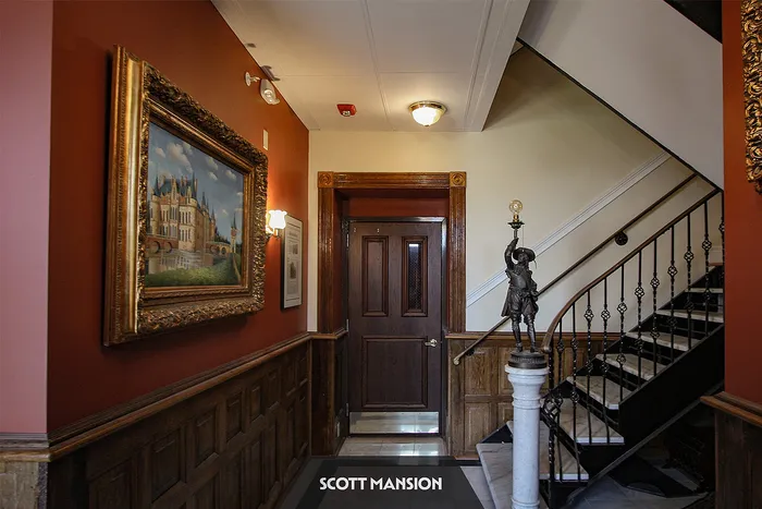 Elegant mansion hallway featuring rich wood accents, classic artwork, and a vintage staircase with intricate railing.