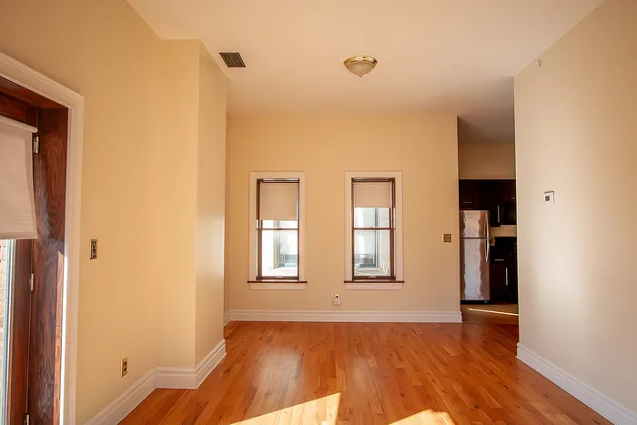 Bright apartment interior featuring polished hardwood floors, large windows, and a glimpse of a modern kitchen with stainless steel appliances.