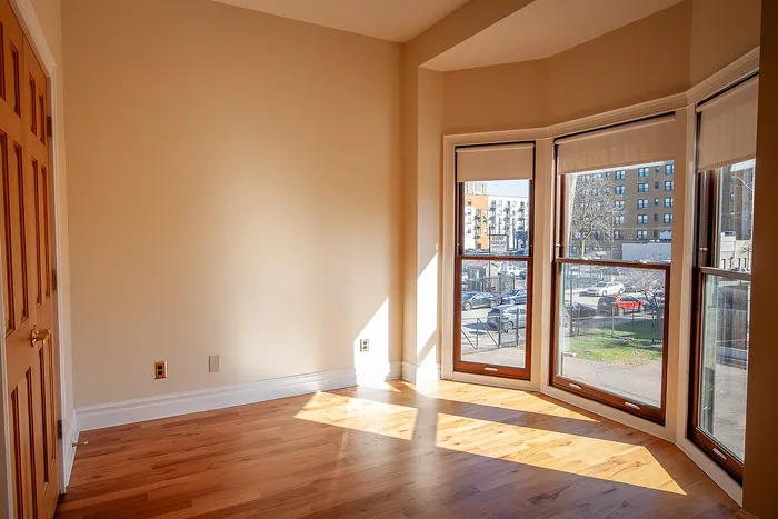 Bright and inviting room with large bay windows, showcasing warm hardwood floors and a sunny urban view.