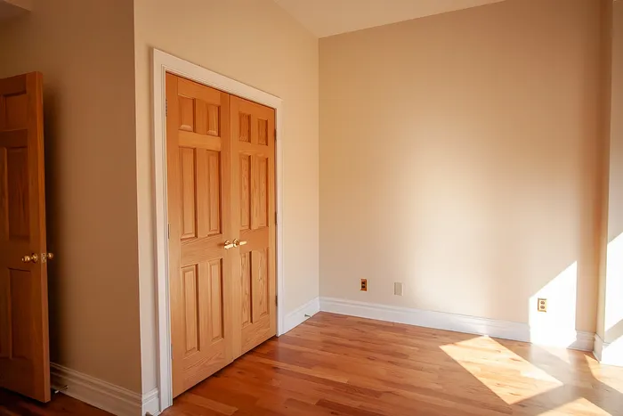 Bright, sunlit room with elegant wooden floors and double wooden closet doors, perfect for a cozy bedroom setting.