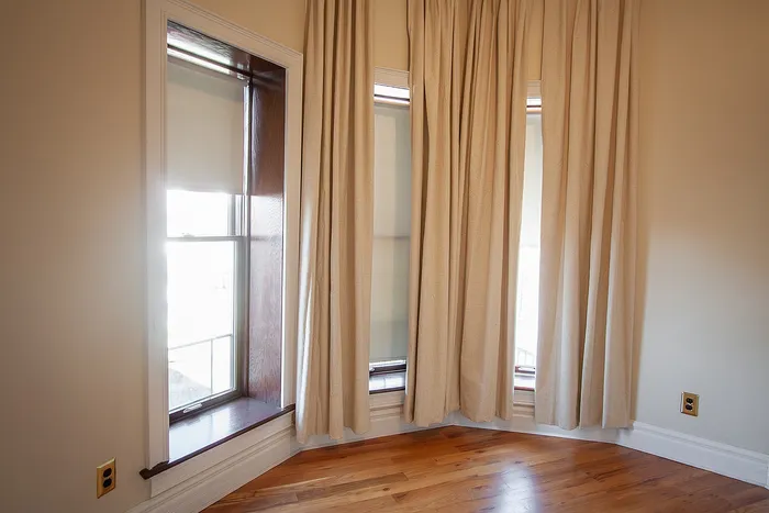 Elegant corner window with abundant natural light, featuring soft beige curtains and polished hardwood floors.